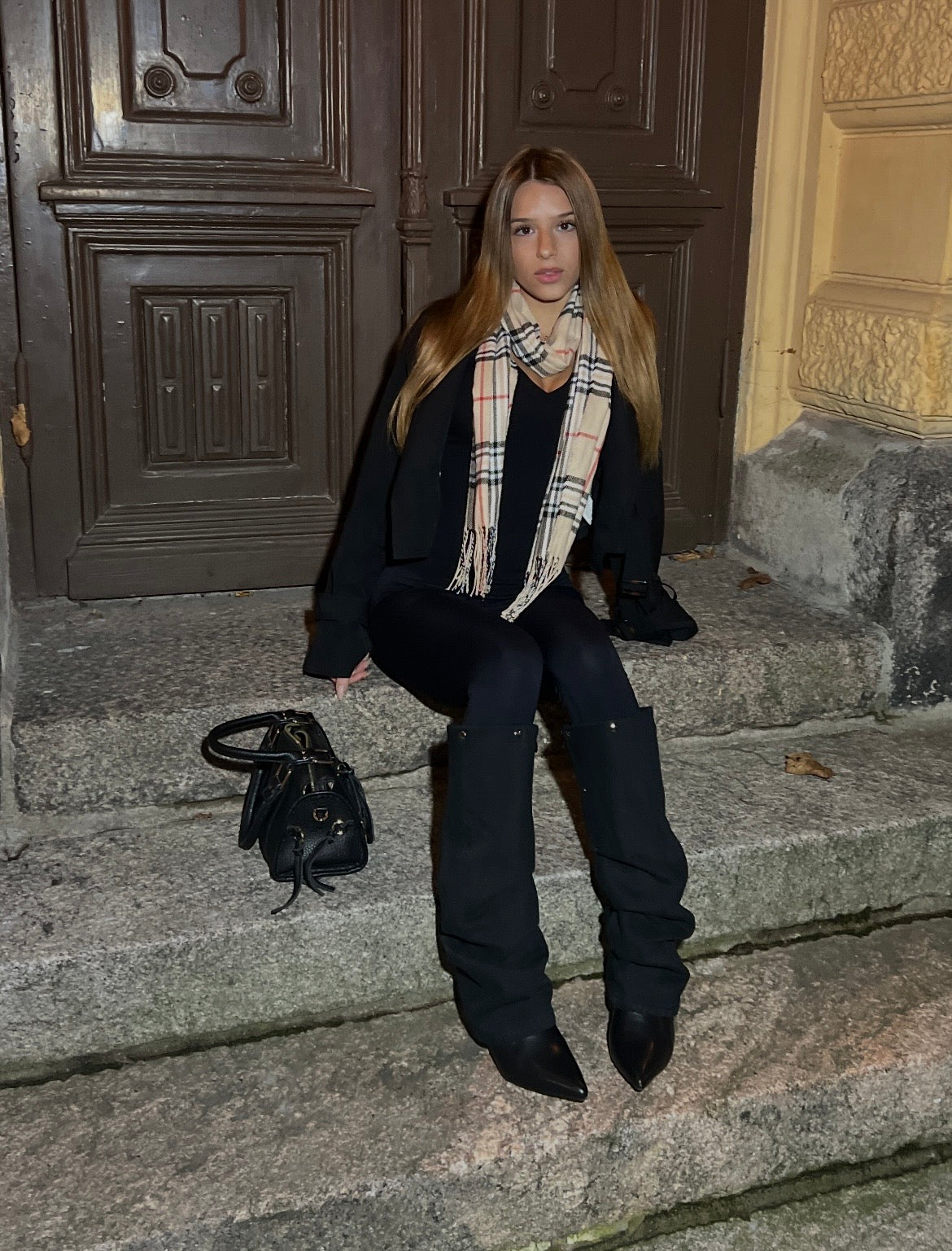 Milena wearing black Denim Heights boots seated on stone steps by a dark wooden door